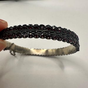 May include: A vintage silver bracelet with two rows of dark red gemstones. The bracelet has an ornate design with a scalloped edge. The gemstones are set closely together, creating a continuous band of colour. The bracelet is likely antique.