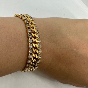 Könnte beinhalten: Ein goldenes Kettenarmband mit klaren Strasssteinen. Das Armband besteht aus einer dicken, massiven Kette mit einem Curb-Link-Design. Die Strasssteine sind in einer Reihe entlang der Ober- und Unterseite der Kette angebracht.