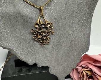 Könnte beinhalten: Eine Vintage-Halskette mit goldfarbenem Anhänger. Der Anhänger zeigt eine Ansammlung von kreisförmigen und trompetenförmigen Elementen. Die Halskette wird auf einer grauen Büste präsentiert, mit einer rosa Rose im Hintergrund. Die Kette ist eine filigrane Gliederkette.