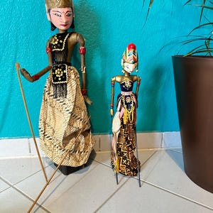 Könnte beinhalten: Zwei traditionelle indonesische Schattenpuppen, eine größer als die andere, mit aufwendigen bemalten Details und kunstvollen Kostümen. Die Puppen sind aus Holz und haben bewegliche Gliedmaßen. Sie stehen auf einem gefliesten Boden.