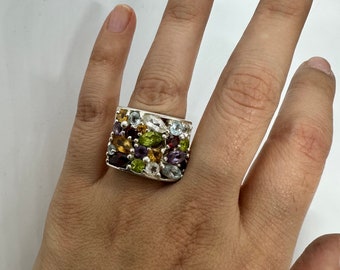 May include: A silver ring with a rectangular face, set with an array of colorful gemstones. The gemstones include shades of purple, green, yellow, and clear stones. The ring is worn on a person's finger.