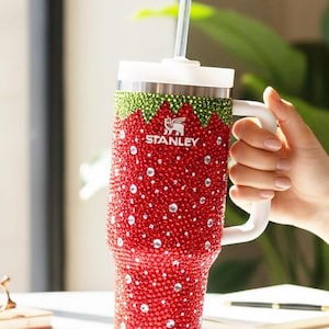 May include: A white ceramic mug with a black handle and the text "But First, Coffee" in a bold, black font. The mug is set against a blurred background, suggesting a cozy setting.