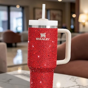 Puede incluir: Un vaso Stanley rojo cubierto de brillantes pedrer&iacute;a. El vaso tiene un asa blanca, una tapa blanca y una pajita blanca. El logotipo de Stanley es visible en la parte delantera. El vaso est&aacute; sobre una mesa de m&aacute;rmol.