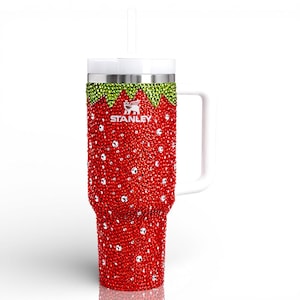 May include: A red and green bedazzled Stanley tumbler with a white handle and straw. The tumbler is covered in red rhinestones, with green rhinestones at the top and a white lid. The Stanley logo is visible.