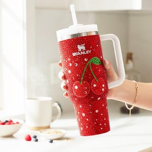 K&ouml;nnte beinhalten: Ein roter Becher mit wei&szlig;em Griff und Deckel, bedeckt mit glitzernden Verzierungen. Der Becher zeigt ein Kirschdesign und das Stanley-Logo. Ein wei&szlig;er Strohhalm ist in den Deckel gesteckt. Der Becher wird in einer Hand gehalten.