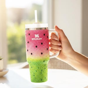 Puede incluir: Un vaso Stanley con tem&aacute;tica de sand&iacute;a con asa y tapa blancas, y una pajita blanca. El vaso est&aacute; decorado con pedrer&iacute;a rosa, verde y negra. El logotipo de Stanley es visible en la parte delantera.