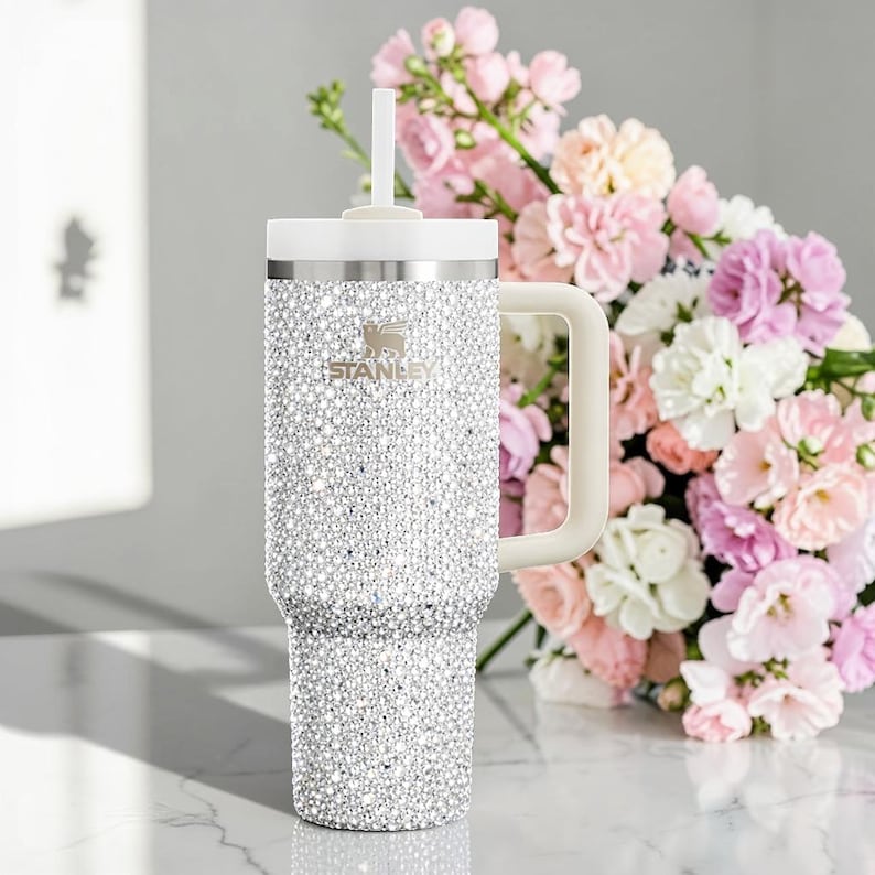 Puede incluir: Un vaso Stanley adornado con pedrer&iacute;a con asa, tapa y pajita blancas. El vaso est&aacute; cubierto de brillantes pedrer&iacute;a. El logotipo de Stanley es visible en la parte delantera. Un ramo de flores rosas, blancas y moradas est&aacute; en el fondo.