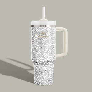 Puede incluir: Un vaso Stanley adornado con pedrer&iacute;a, con asa, tapa y pajita blancas. El vaso de acero inoxidable est&aacute; cubierto de brillantes pedrer&iacute;a. El logotipo de Stanley es visible. Ideal para bebidas fr&iacute;as o calientes.