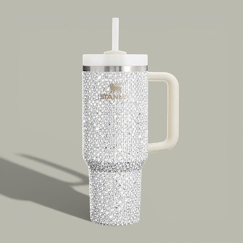 Puede incluir: Un vaso Stanley adornado con pedrer&iacute;a, con asa, tapa y pajita blancas. El vaso de acero inoxidable est&aacute; cubierto de brillantes pedrer&iacute;a. El logotipo de Stanley es visible. Ideal para bebidas fr&iacute;as o calientes.