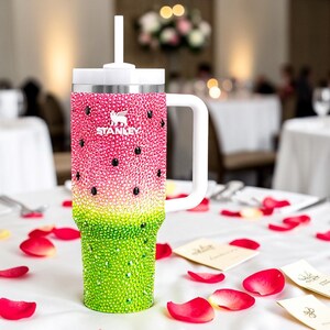 Puede incluir: Un vaso Stanley adornado con pedrer&iacute;a con un dise&ntilde;o de sand&iacute;a. El vaso presenta un degradado rosa y verde, con detalles en negro. Tiene un asa, tapa y pajita blancos. El logotipo de Stanley es visible. Sobre un mantel blanco con p&eacute;talos de rosa.