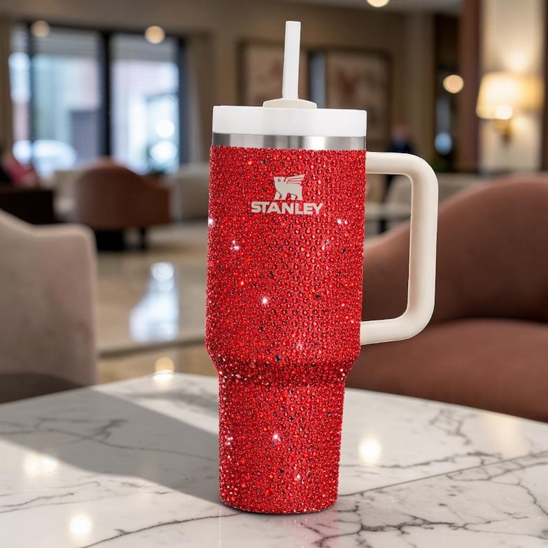 Puede incluir: Un vaso Stanley rojo cubierto de brillantes pedrer&iacute;a. El vaso tiene un asa blanca, una tapa blanca y una pajita blanca. El logotipo de Stanley es visible en la parte delantera. El vaso est&aacute; sobre una mesa de m&aacute;rmol.