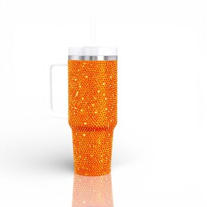 May include: An orange, bedazzled tumbler with a white handle, lid, and straw. The tumbler is covered in small, sparkling orange rhinestones. The handle is rectangular and attached to the side of the tumbler. The straw is white and straight.