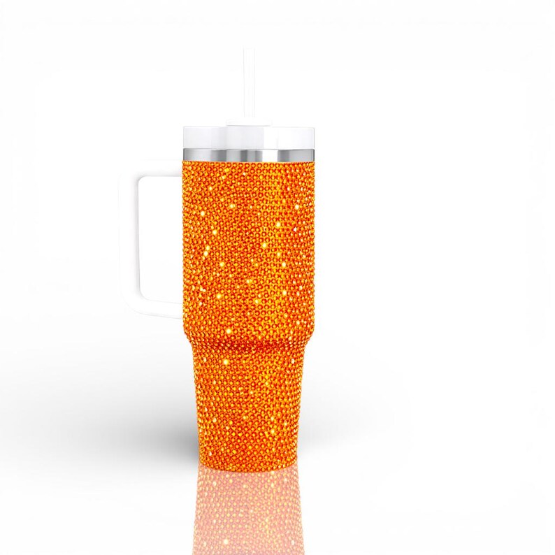 May include: An orange, bedazzled tumbler with a white handle, lid, and straw. The tumbler is covered in small, sparkling orange rhinestones. The handle is rectangular and attached to the side of the tumbler. The straw is white and straight.