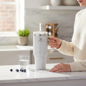 Puede incluir: Un vaso Stanley grande, adornado con pedrer&iacute;a, con asa y tapa blancas, y una pajita blanca. El vaso est&aacute; cubierto de peque&ntilde;os y brillantes pedrer&iacute;a. Un vaso de agua y ar&aacute;ndanos est&aacute;n en el mostrador.