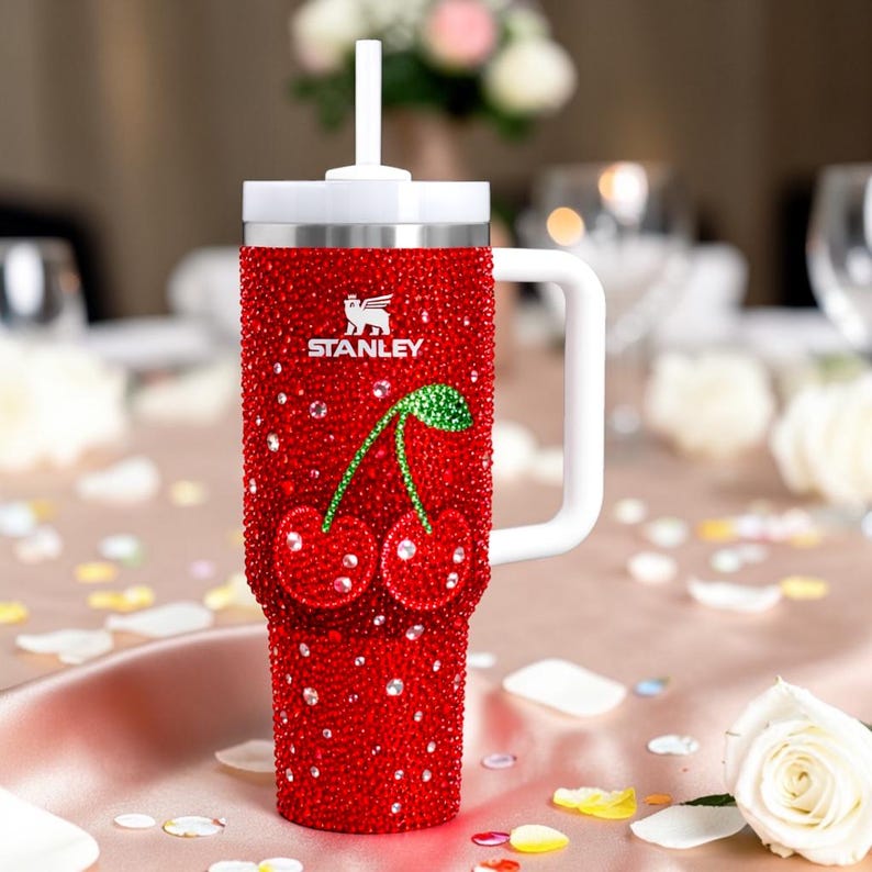 K&ouml;nnte beinhalten: Roter Stanley-Becher mit Strasssteinen und Kirschdesign. Er hat einen wei&szlig;en Griff, Deckel und Strohhalm. Das Stanley-Logo ist sichtbar. Der Becher steht auf einem Tisch mit Bl&uuml;tenbl&auml;ttern.