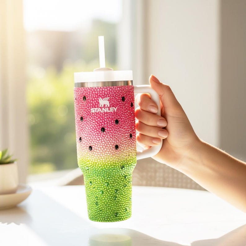 Puede incluir: Un vaso Stanley con tem&aacute;tica de sand&iacute;a con asa y tapa blancas, y una pajita blanca. El vaso est&aacute; decorado con pedrer&iacute;a rosa, verde y negra. El logotipo de Stanley es visible en la parte delantera.