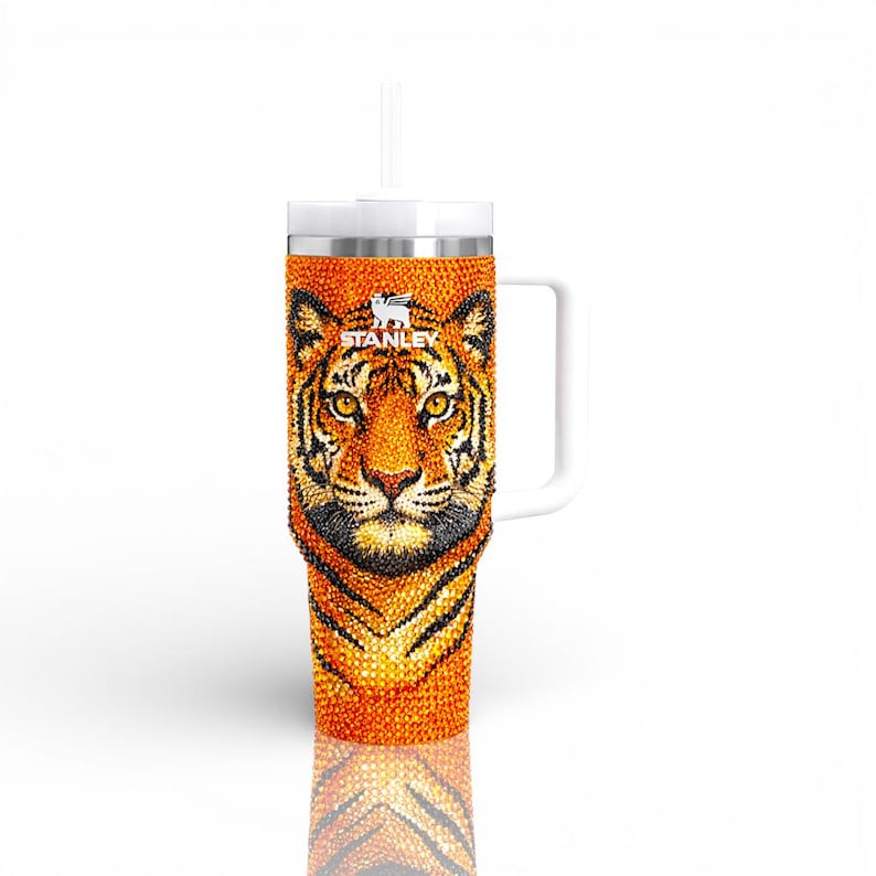 May include: A Stanley tumbler with a tiger design. The orange and black tiger face is embellished with rhinestones. The tumbler has a white handle and a white straw. The Stanley logo is visible on the top.