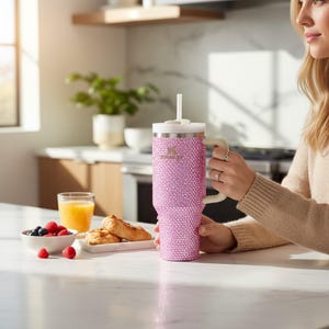 K&ouml;nnte beinhalten: Ein pinkfarbener, mit Strass besetzter Stanley-Becher mit wei&szlig;em Griff und Deckel sowie einem wei&szlig;en Strohhalm. Der Becher wird von einer Person in einer K&uuml;che gehalten, mit einem Fr&uuml;hst&uuml;ck aus Obst und Geb&auml;ck in der N&auml;he.