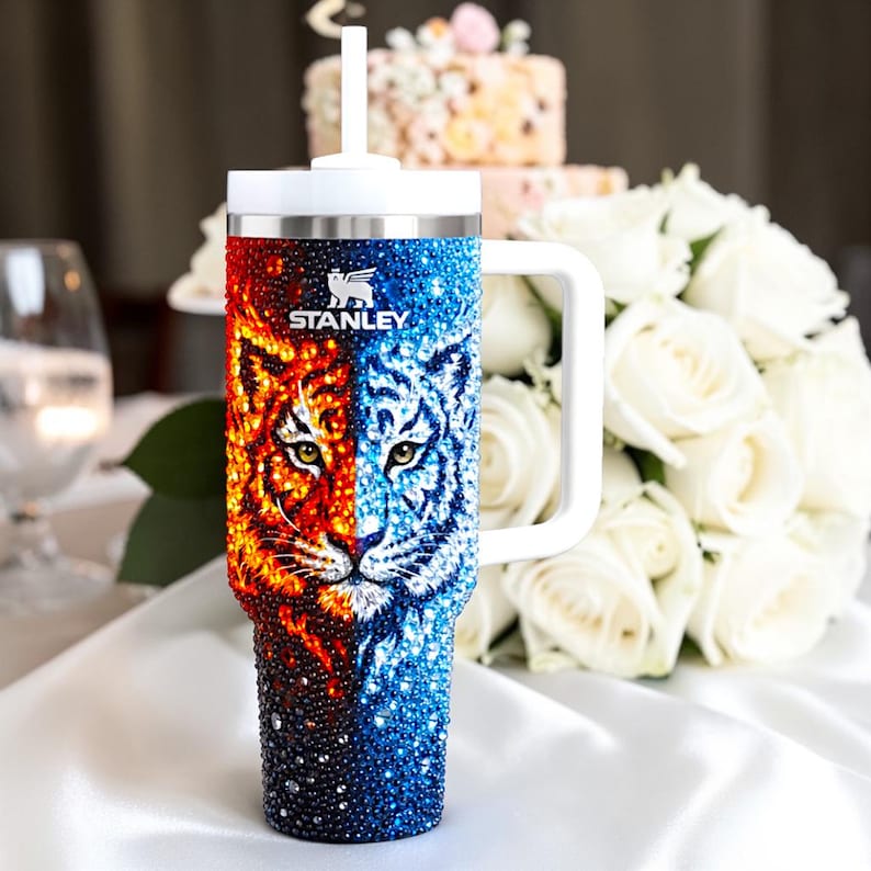 Puede incluir: Un vaso Stanley de acero inoxidable con asa y tapa blancas. El vaso presenta un dise&ntilde;o dividido, con una cara de tigre en rojo y naranja por un lado y azul y blanco por el otro. El logotipo de Stanley es visible.