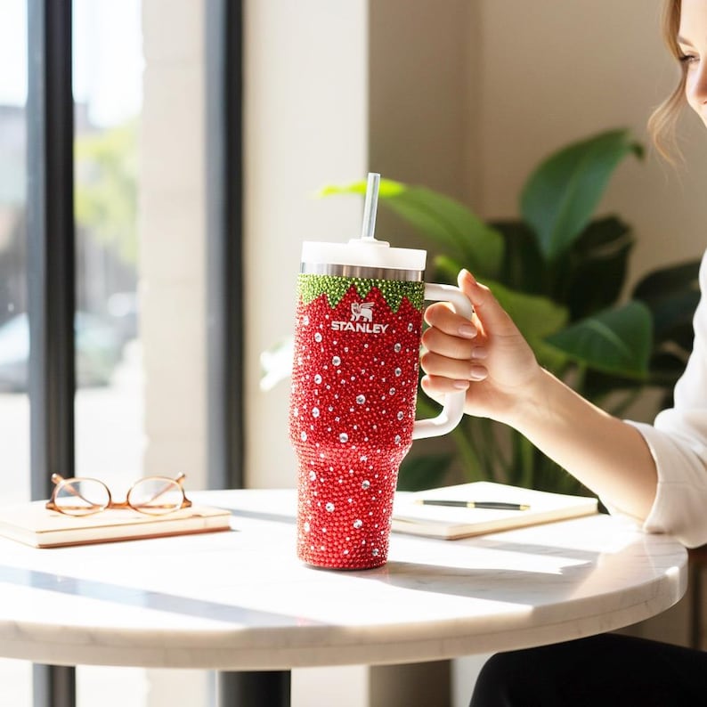 May include: A white ceramic mug with a black handle and the text "But First, Coffee" in a bold, black font. The mug is set against a blurred background, suggesting a cozy setting.