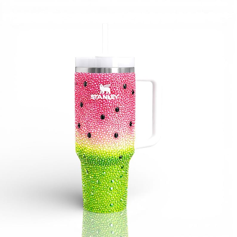 Puede incluir: Un vaso de la marca Stanley con dise&ntilde;o de sand&iacute;a. La mitad superior es rosa con acentos de semillas negras, y la mitad inferior es verde. El vaso est&aacute; cubierto de brillantes pedrer&iacute;a y tiene un asa y una pajita blancas.