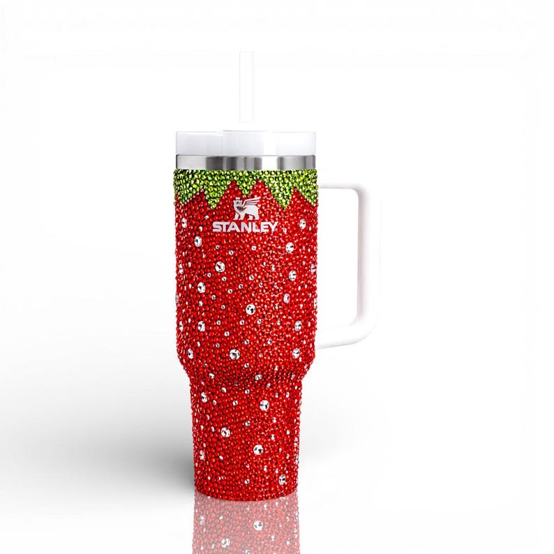 May include: A red and green bedazzled Stanley tumbler with a white handle and straw. The tumbler is covered in red rhinestones, with green rhinestones at the top and a white lid. The Stanley logo is visible.