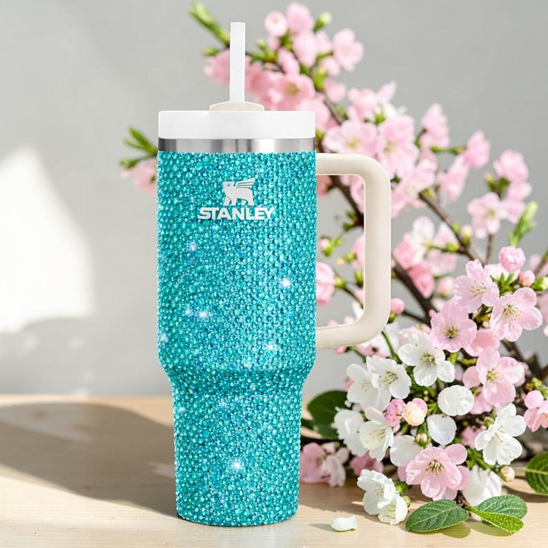 Puede incluir: Un vaso Stanley turquesa con un exterior brillante, similar a una joya. Cuenta con un asa, tapa y pajita blancos. El logotipo de Stanley es visible. El fondo incluye flores rosas y blancas.