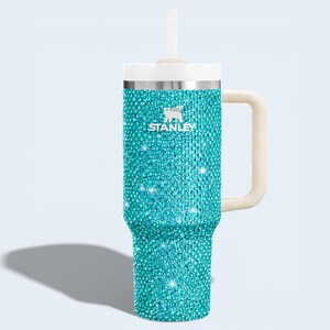 Puede incluir: Un vaso Stanley turquesa con un exterior adornado con pedrer&iacute;a. Tiene una tapa y pajita blancas, un asa color crema y el logotipo de Stanley. Dise&ntilde;ado para bebidas fr&iacute;as o calientes.