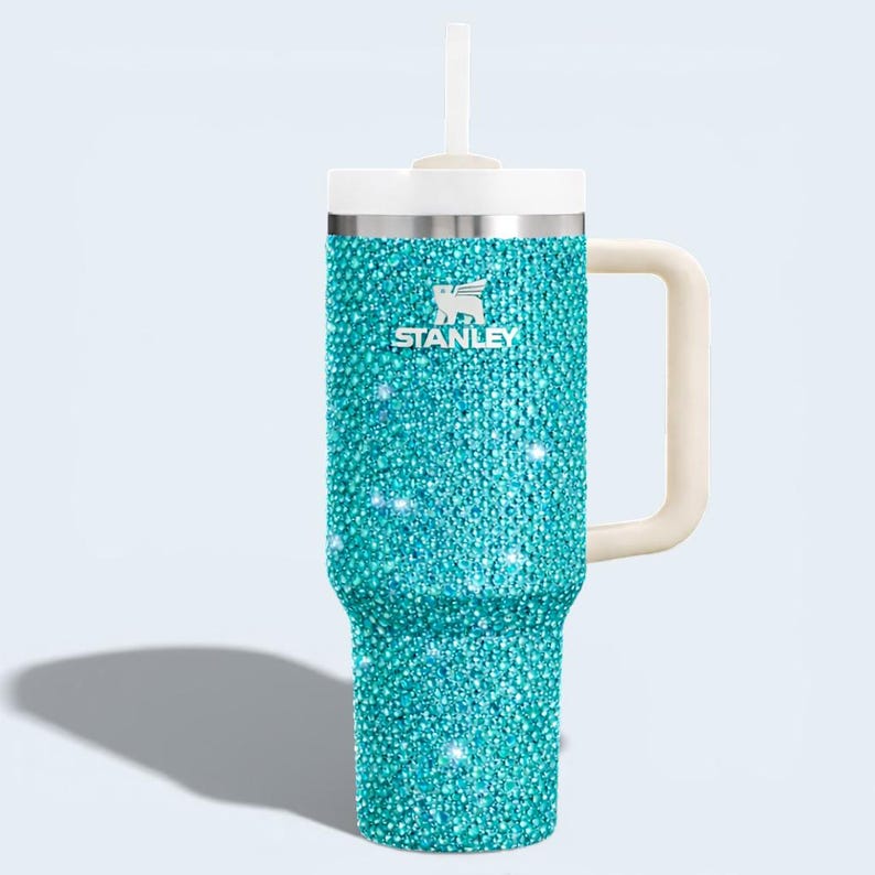Puede incluir: Un vaso Stanley turquesa con un exterior adornado con pedrer&iacute;a. Tiene una tapa y pajita blancas, un asa color crema y el logotipo de Stanley. Dise&ntilde;ado para bebidas fr&iacute;as o calientes.