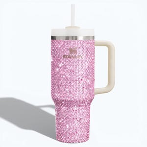 K&ouml;nnte beinhalten: Ein pinkfarbener, mit Strasssteinen besetzter Stanley-Becher mit wei&szlig;em Deckel, Griff und Strohhalm. Der Becher tr&auml;gt das Stanley-Logo und ist mit glitzernden, pinkfarbenen Strasssteinen bedeckt. Der Griff ist hellbeige.