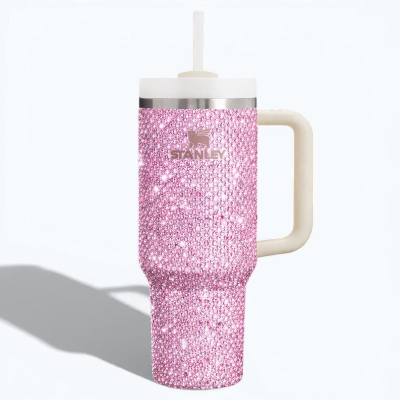 K&ouml;nnte beinhalten: Ein pinkfarbener, mit Strasssteinen besetzter Stanley-Becher mit wei&szlig;em Deckel, Griff und Strohhalm. Der Becher tr&auml;gt das Stanley-Logo und ist mit glitzernden, pinkfarbenen Strasssteinen bedeckt. Der Griff ist hellbeige.