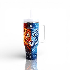 Puede incluir: Un vaso Stanley de acero inoxidable con asa y pajita blancas. El vaso presenta un dise&ntilde;o dividido con la cara de un tigre, la mitad en naranja y rojo fuego, la otra mitad en tonos azules fr&iacute;os. El logotipo de Stanley es visible.
