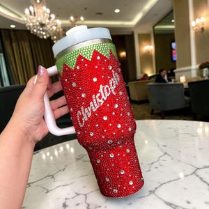 May include: A red tumbler covered in rhinestones, with a green and silver trim. The tumbler has a white handle and lid. The name "Christopher" is written in silver script on the side. The tumbler is designed to hold cold beverages.