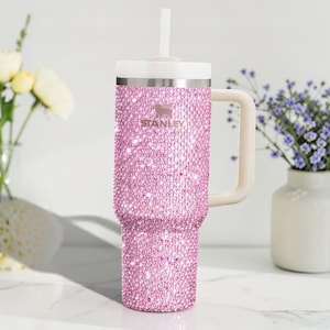K&ouml;nnte beinhalten: Ein pinkfarbener, mit Strass besetzter Stanley-Becher mit wei&szlig;em Griff, Deckel und Strohhalm. Der Becher ist mit kleinen, funkelnden, rosa Strasssteinen bedeckt. Das Stanley-Logo ist auf der Vorderseite sichtbar.