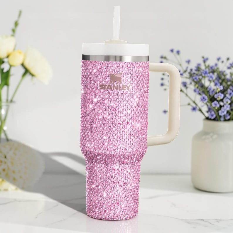 K&ouml;nnte beinhalten: Ein pinkfarbener, mit Strass besetzter Stanley-Becher mit wei&szlig;em Griff, Deckel und Strohhalm. Der Becher ist mit kleinen, funkelnden, rosa Strasssteinen bedeckt. Das Stanley-Logo ist auf der Vorderseite sichtbar.