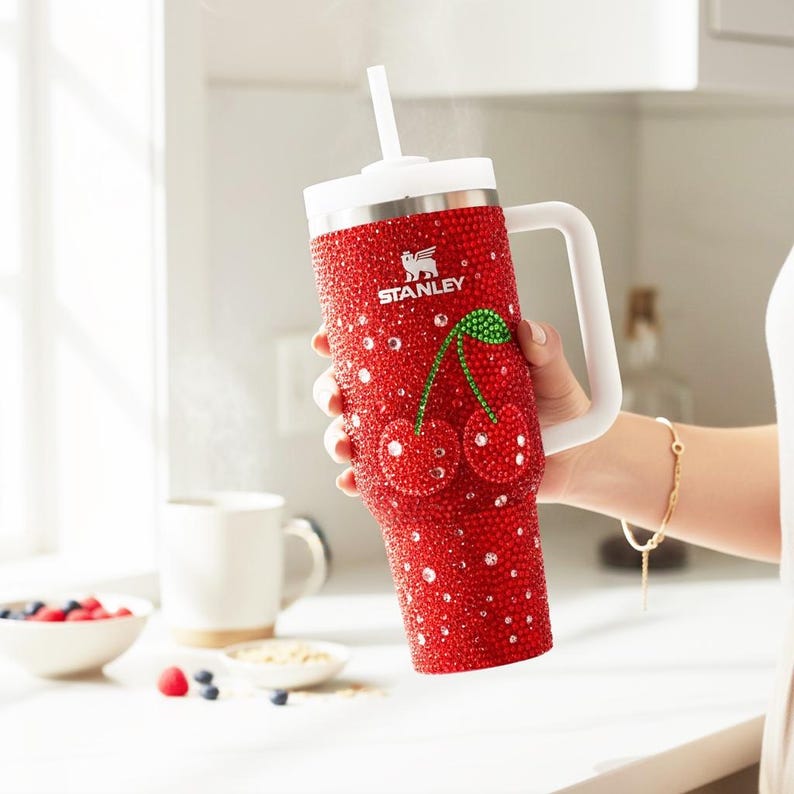 K&ouml;nnte beinhalten: Ein roter Becher mit wei&szlig;em Griff und Deckel, bedeckt mit glitzernden Verzierungen. Der Becher zeigt ein Kirschdesign und das Stanley-Logo. Ein wei&szlig;er Strohhalm ist in den Deckel gesteckt. Der Becher wird in einer Hand gehalten.
