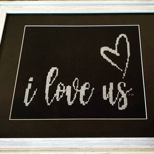 Può includere: Un ricamo a punto croce incorniciato con sfondo nero e ricamo bianco. Il ricamo dice "i love us" con un cuore sopra le parole.