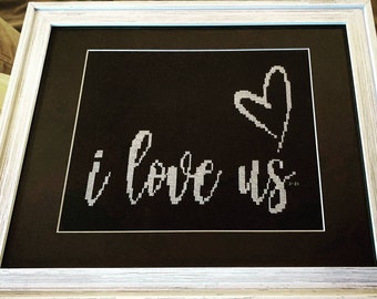 Könnte beinhalten: Ein gerahmtes Kreuzstichbild mit schwarzem Hintergrund und weißer Stickerei. Die Stickerei sagt "i love us" mit einem Herzen über den Worten.