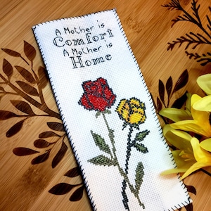 Puede incluir: Un marcapáginas de punto de cruz blanco con un diseño de rosa roja y amarilla y el texto "Una madre es consuelo, una madre es hogar".