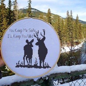 May include: A cross-stitch embroidery hoop featuring a deer and a buck silhouette with the text "You Keep Me Safe, I'll Keep You Wild." The design is in black on a white background, set against a scenic outdoor backdrop.