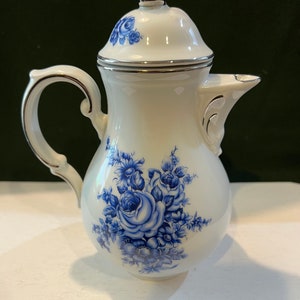 May include: A white porcelain creamer with a silver lid and blue floral design. The creamer has a rounded body and a curved handle.