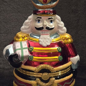 May include: A decorative Christmas ornament shaped like a nutcracker soldier. The ornament features a red jacket with gold trim, a gold glitter hat, and a white face with a black mustache. It holds a gift box with a green cross.