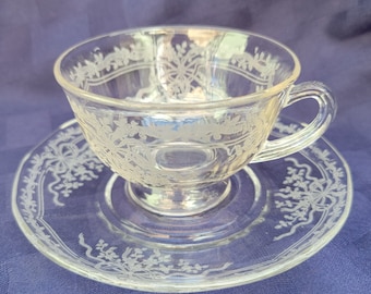 May include: A clear glass teacup and saucer set with an etched floral design. The cup has a handle and a wide, shallow bowl. The saucer is round and has a scalloped edge.