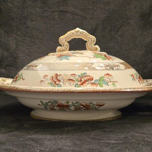 May include: Cream-colored, lidded serving dish with floral and geometric patterns. The dish features a handle on the lid and side handles. The floral design includes red, orange, and green accents. The dish is likely made of ceramic or porcelain.