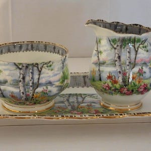 May include: A porcelain creamer, sugar bowl, and rectangular serving tray set featuring a painted landscape of birch trees, colorful flowers, and a lake. Each piece has a white base, gold-trimmed scalloped edges, and a dark gray border pattern.
