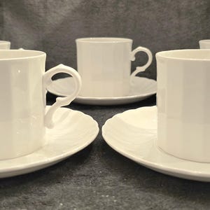 May include: A set of five white porcelain teacups and saucers. The cups have a square shape and ornate handles. The saucers have a scalloped edge. The set is arranged on a dark gray surface.