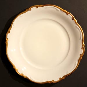 May include: A round white porcelain plate featuring a scalloped edge with a metallic gold trim. The center of the plate is smooth and plain, while the rim displays a raised, embossed relief pattern that follows the curves of the scalloped border.