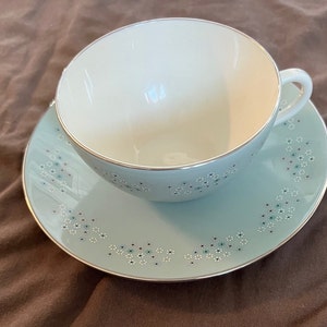 May include: A vintage teacup and saucer set. The cup and saucer are a light blue with a silver trim. The saucer and cup are decorated with small floral patterns in white, black, and teal. The inside of the cup is white.
