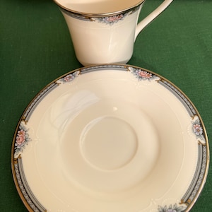 May include: A white porcelain teacup and saucer with a gold and blue trim and floral design. The saucer has a scalloped edge.
