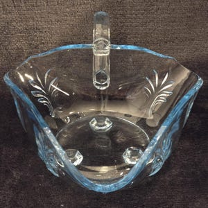 May include: A clear, light blue glass serving dish with a handle. The dish has a scalloped edge and is decorated with etched floral designs. The base has three small feet. The handle is clear and has a decorative design.